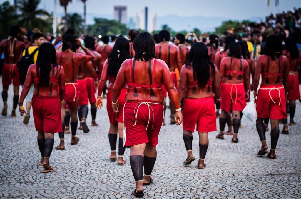 Grupo de indígenas com roupas e o corpo pintados de vermelho caminha numa rua em manifestação.