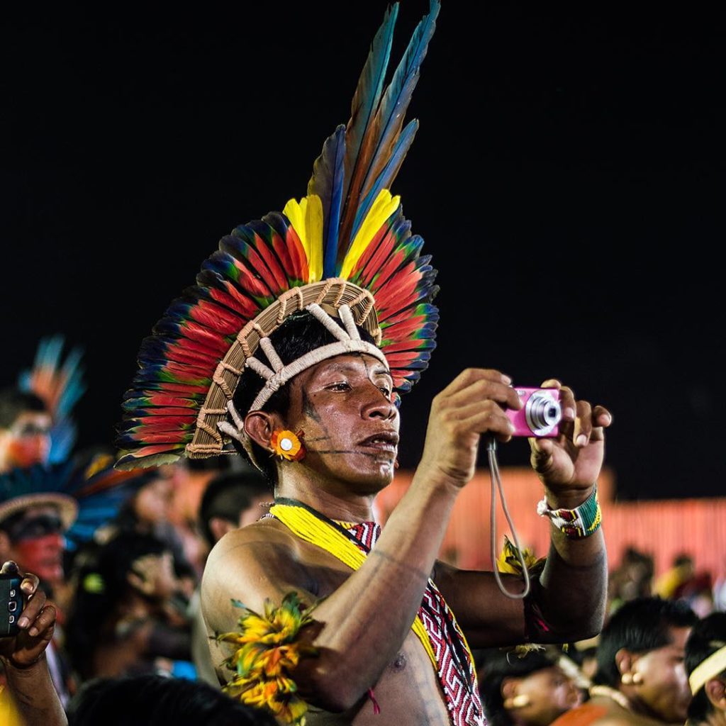 Homem indígena com roupas tradicionais e cocar multicolorido fotografa em meio a uma multidão
