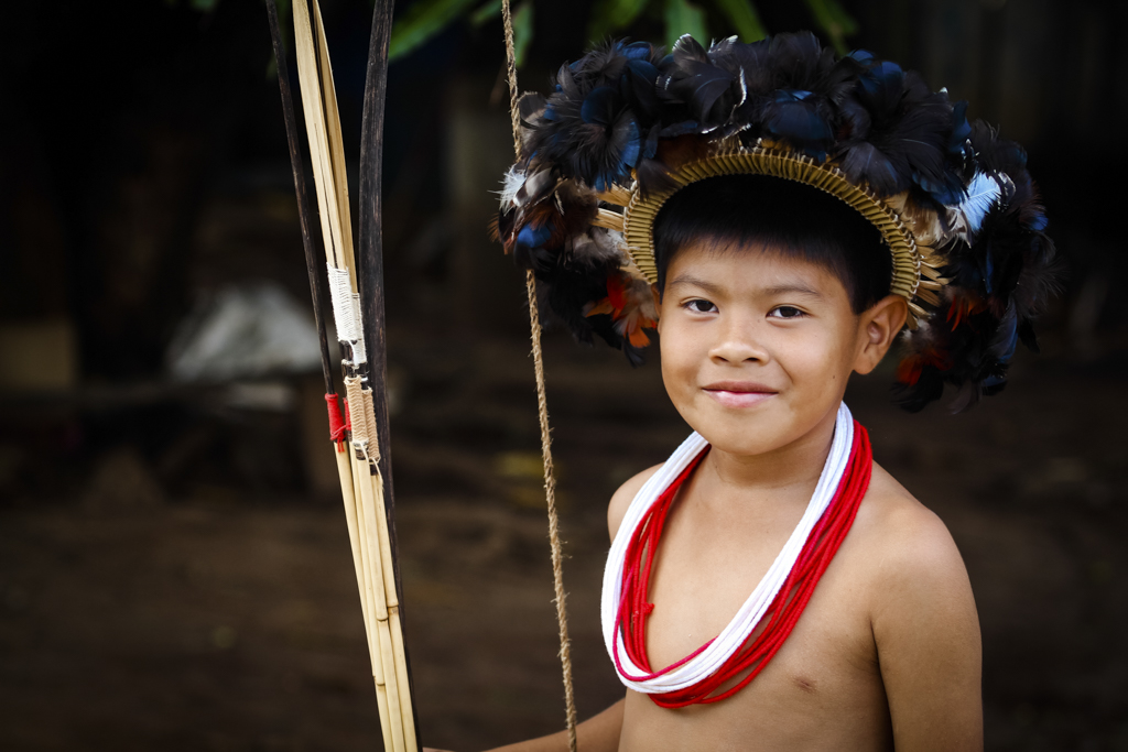 Criança indígena com arco e colares em meio a uma aldeia.