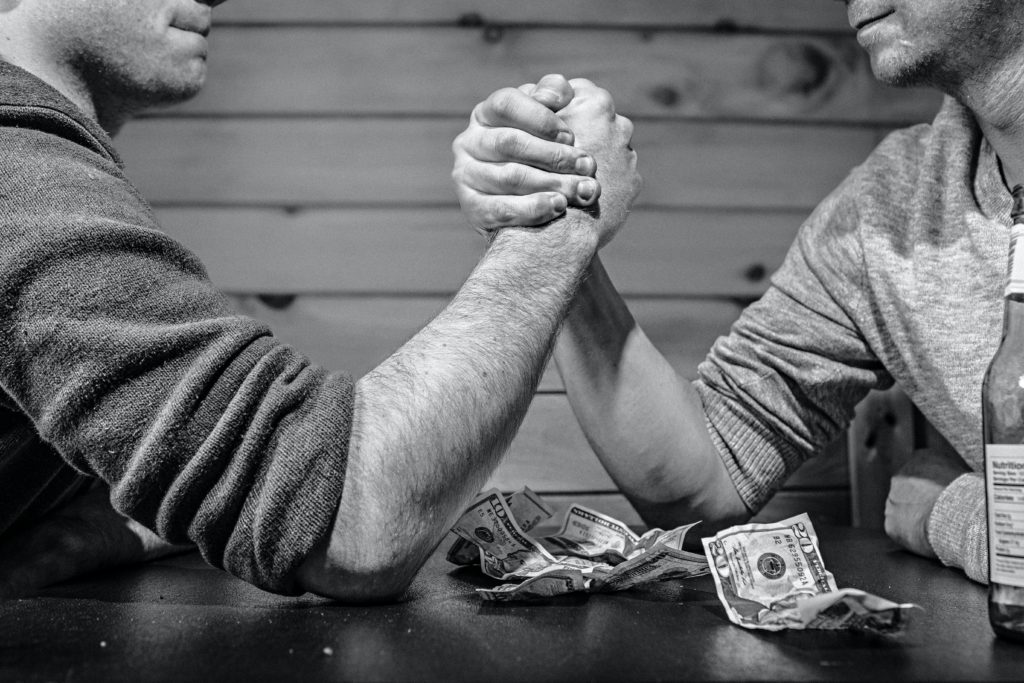 Foto preto e branca com dois homens jogando queda de braço numa mesa com notas de dinheiro sobre ela, ilustrando de forma metafórica os percalços do diálogo quando entendido como competição.
