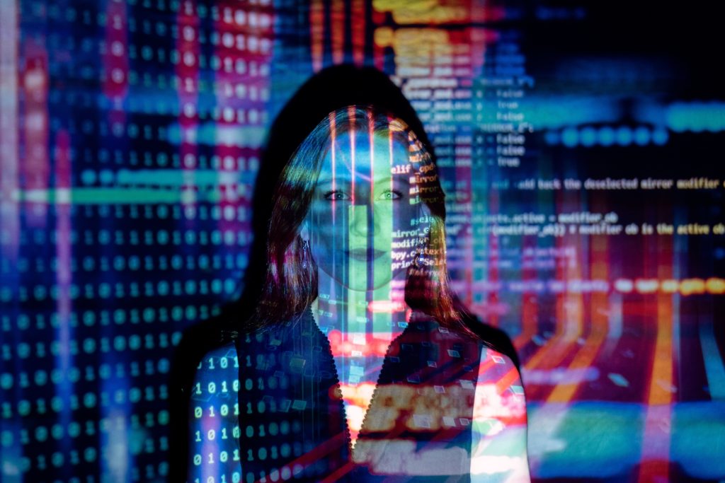 Foto de uma mulher com projeções de código em diversas cores sobre ela e sobre a parede ao fundo da foto, em menção à era dos algoritmos