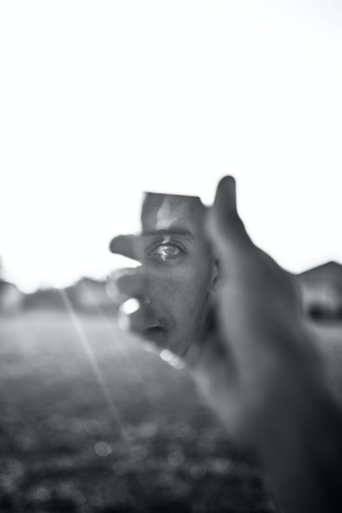 foto preto e branca de uma mão segurando um espelho quebrado que reflete um rosto de um homem.