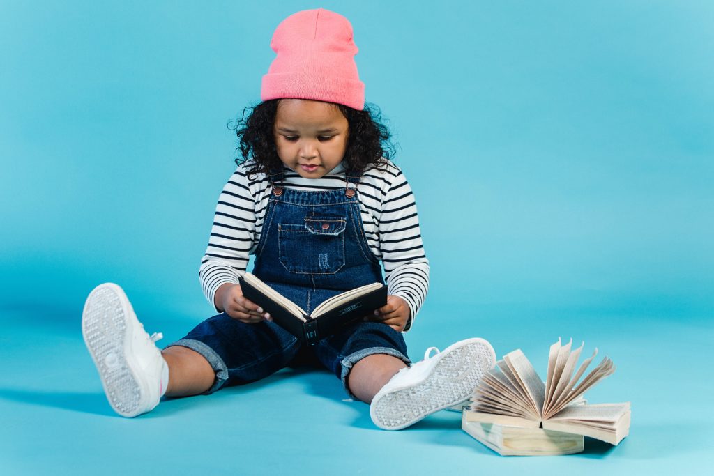 Fundo azul com uma menina negra de tca rosa lendo um livro no chão.