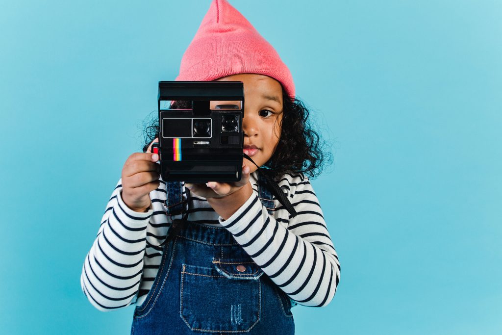 Fundo azul com menina negra fotografando com uma câmera polaroid, simbolizando o tema do artigo sobre o poder da infância