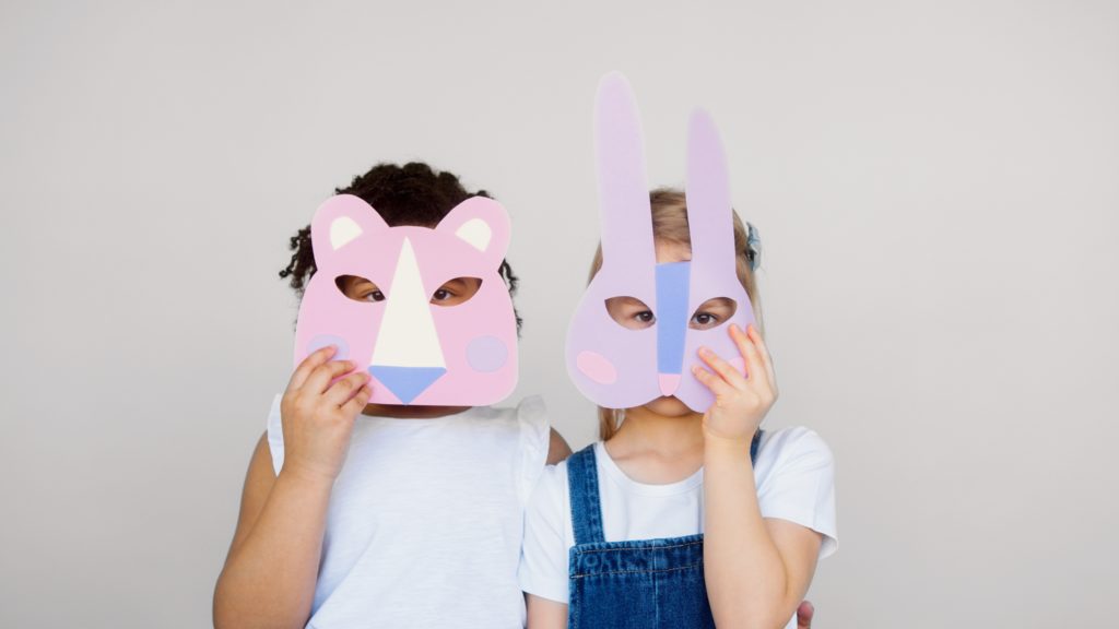Fundo branco com duas crianças, uma branca e outra negra usando máscaras de papel em tons de rosa e azul claros em referência ao tema das histórias de ninar.