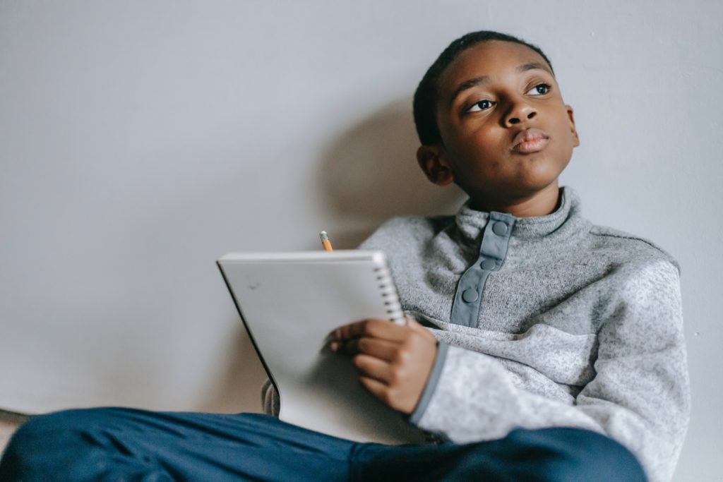 Menino negro sentado na cama escrevendo em um caderno em referência ao tema das histórias de ninar.