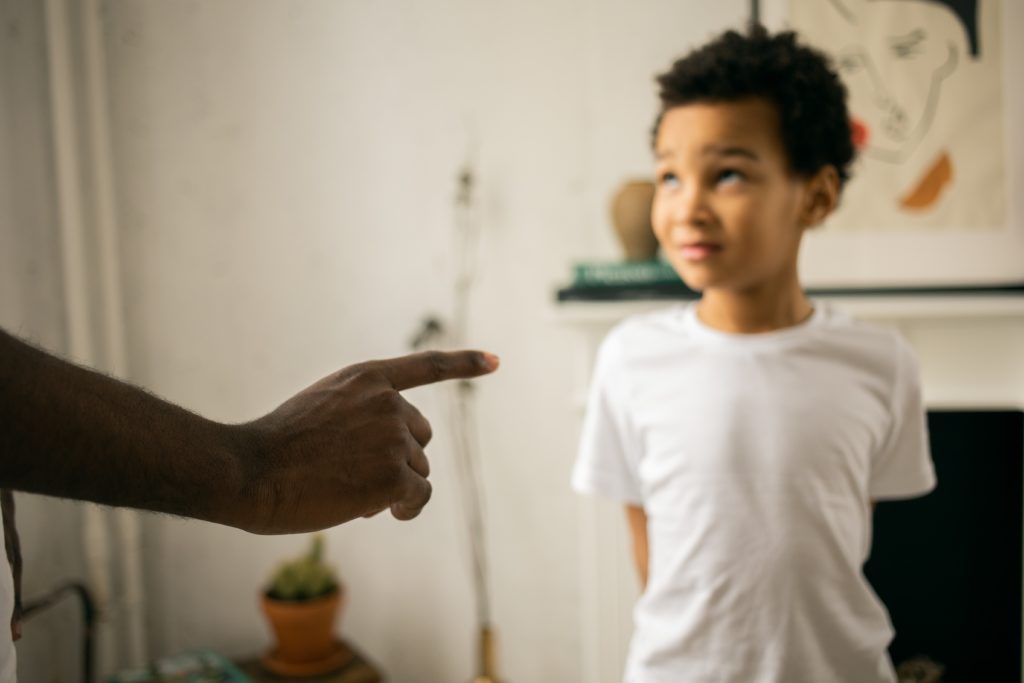 Menino negro com cara de constrangimento e uma mão adulta apontando o dedo para ele numa relação de parentalidade.