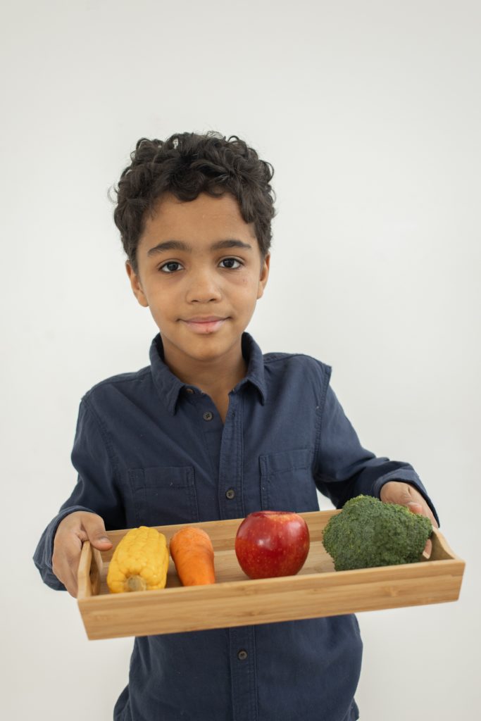 Menino negro segurando uma bandeja com frutas e legumes. Fundo branco.